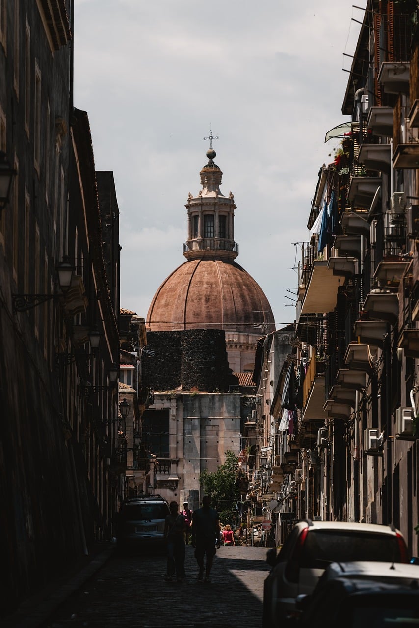 Catania, Kathedrale, Wahrzeichen der Stadt, enge Gasse, historische Architektur, bedeckter Himmel, italienische Stadtansicht, Kopfsteinpflaster, Fußgänger, alte Gebäude, Süditalien, Reisefotografie, Tourismus, städtische Silhouette, Stadtleben, Kuppel, Straßenszene, lokaler Architekt
