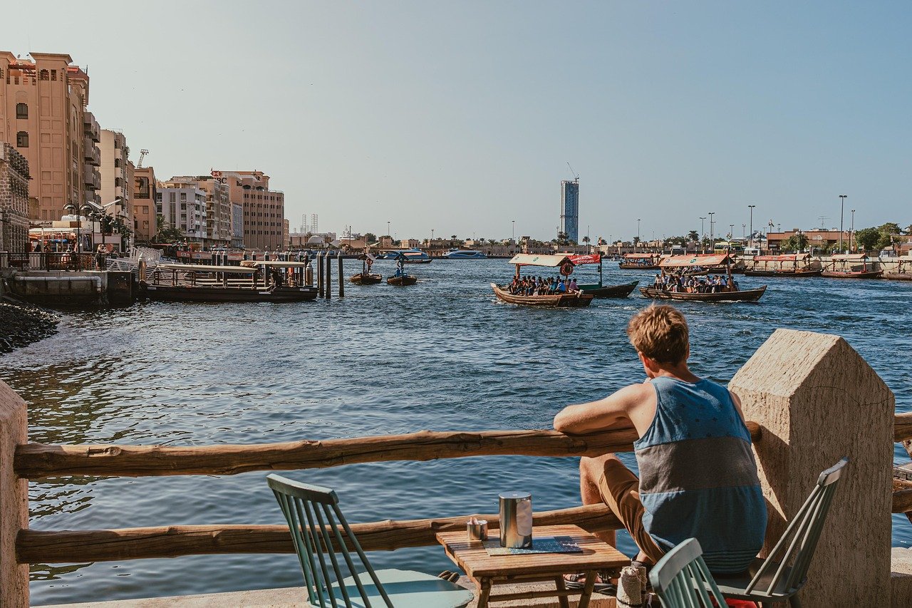 Dubai Creek, Uferpromenade, sitzender Mann, Abra-Boote, traditionelle Boote, Natur, Dubai Wassertaxis, entspannende Aussicht, Außensitzplätze, Creek-Aussicht, Menschen, Abra-Fahrt, sonniger Tag, Sightseeing, Ufercafé, Bootsfahrt, lässige Szene, Stadtbild, städtische Aussicht, Uferpromenade
